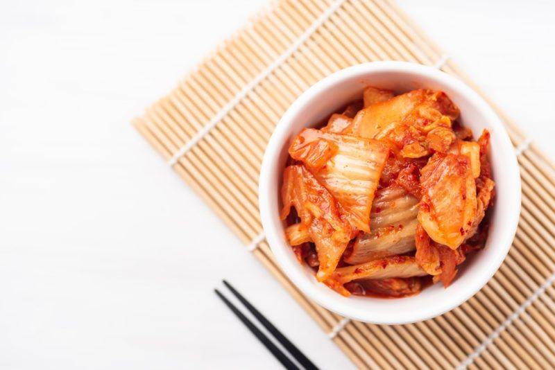 A white bowl of kimchi, next to chopsticks on a bamboo mat