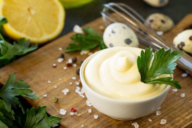 A small white dish containing homemade mayonnaise next to salt, pepper, a lemon half, and herbs