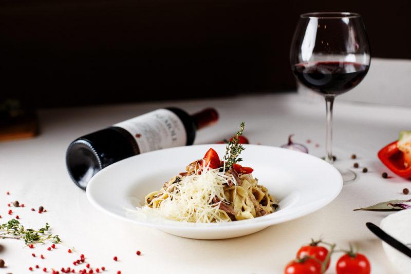 A white dish on a table with pasta, next to a bottle of red wine and a glass of the wine