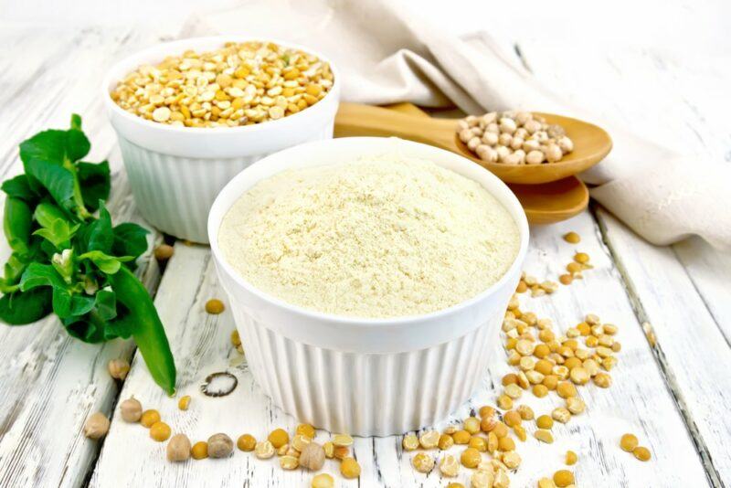 A white wooden table with a white bowl of pea protein powder and a smaller one of split yellow peas