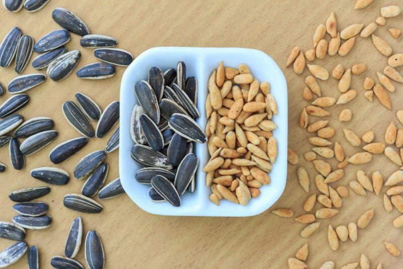 A white segmented dish, where half contains shelled sunflower seeds and the other half has unshelled ones, with more of each scattered on the table