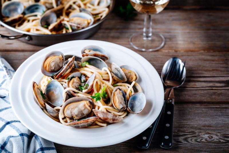 A white dish with spaghetti alle vongole next to a pan of the pasta and a glass of wine