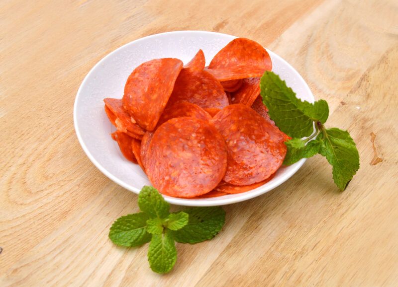 A small white dish of turkey pepperoni on a wooden board with a few pieces of leaves