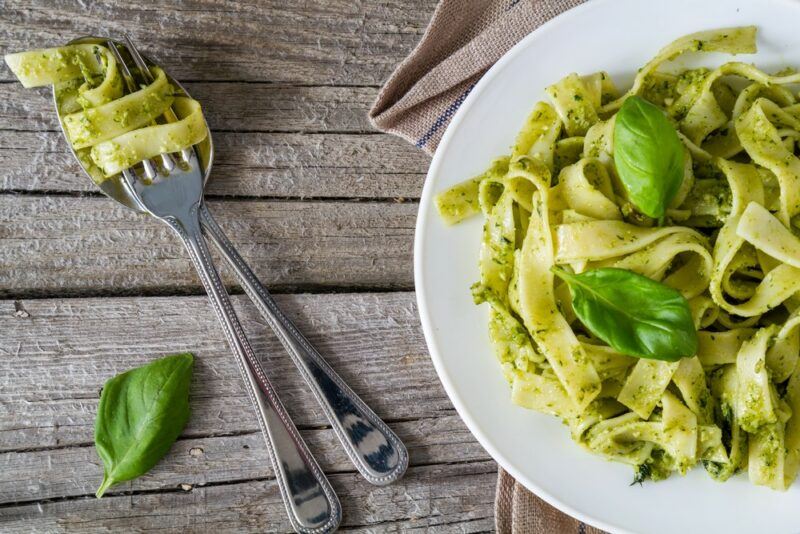 A white dish with pesto pasta next to a knife and fork with pasta curled around them