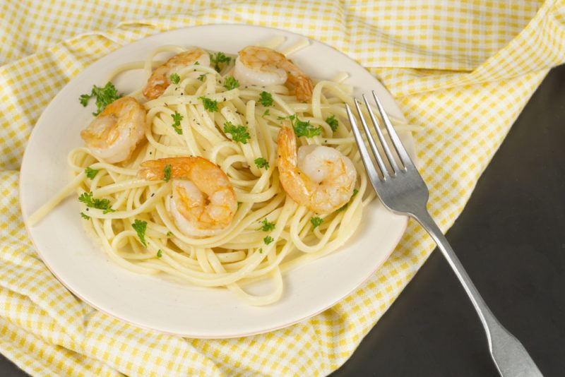 A white plate on a light cloth with shrimp scampi next to a fork