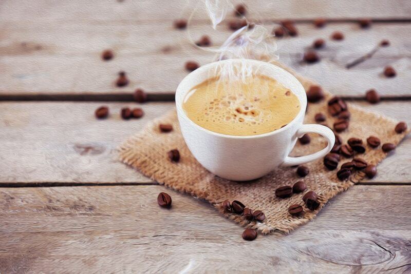 A wooden table with a white mug of decaf coffee that is surrounded by many coffee beans