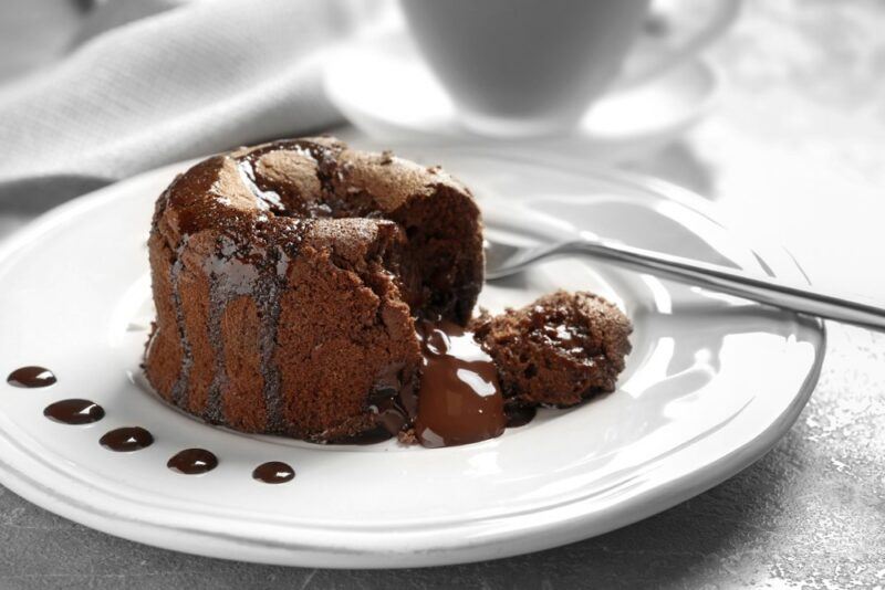 A white plate containing a chocolate lava cake with a spoon and a few dots of chocolate sauce around the edge