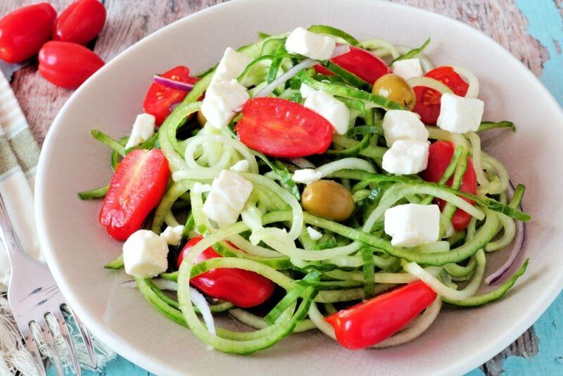 A white plate with a zoodle, feta, olive, and tomato salad