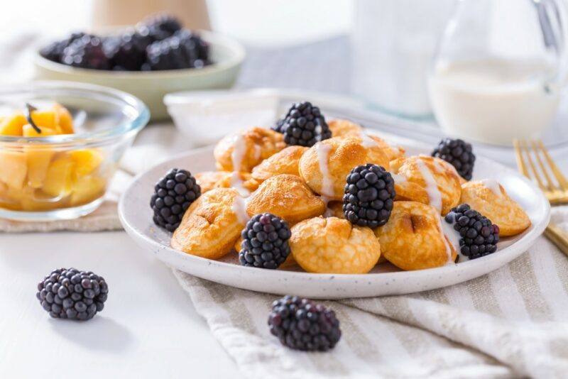 A white table with a cloth and a white plate containing small Dutch pancakes and blackberries, with a dish of mango and another of blackberries in the background