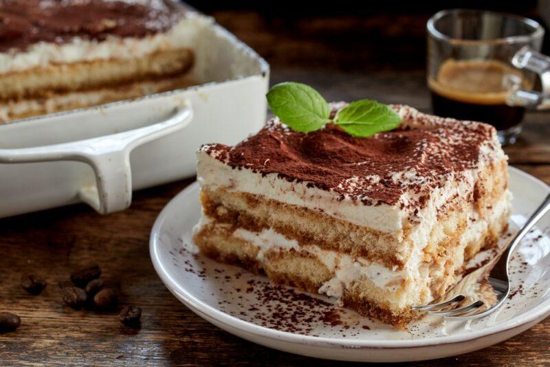 A large white dish containing tiramisu, next to a plate with a square of tiramisu. There's a glass mug of coffee in the background