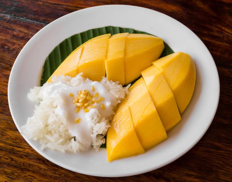 A white plate of kao niew ma muang, which includes two sliced mangos, a mango leaf, rice, and condensed milk