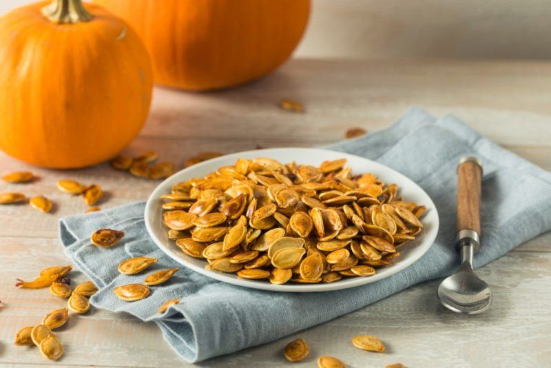 A white plate of pumpkin seeds in front of a large pumpkin and a small one