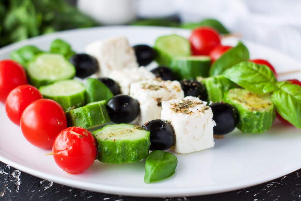 A white plate with four Greek salad skewers, each includes cherry tomatoes, cucumber, feta, olives, and herbs