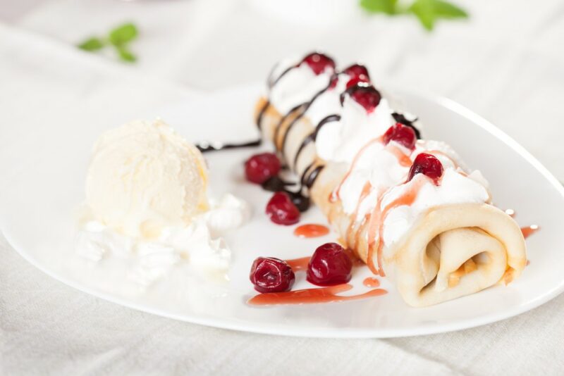 A white plate with a rolled crepe that was part of a dessert called firewood on snow, next to a scoop of ice cream