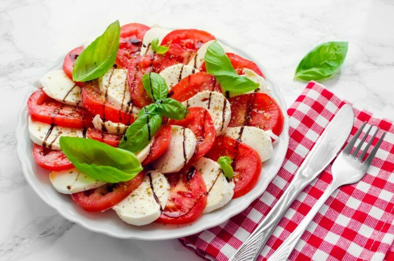 A white plate containing a large caprese salad, complete with balsamic vinegar, with a knife and fork next to it