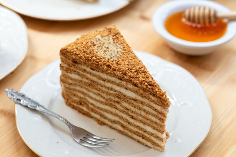 A white plate with a layered medovik cake on a plate, next to a fork, with a small tray of honey in the background