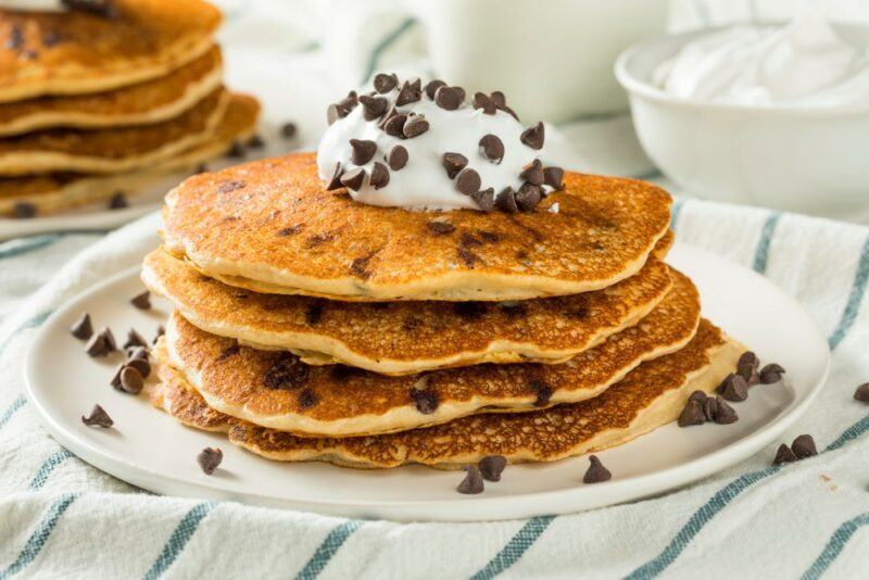 A white plate with a pile of four chocolate chip pancakes. The stack is topped with white sauce and plenty of chocolate chips. There is another stack of pancakes out of focus in the background, plus a white bowl of more icing.