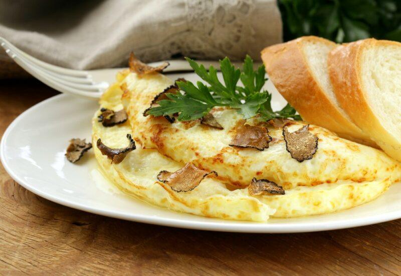 A wooden table with a white plate that has an omelet and sliced truffles, next to sliced bread