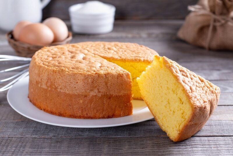 A white plate with a castella cake, where a large slice has been cut out of it. There are eggs, a sugar container, and other items in the background