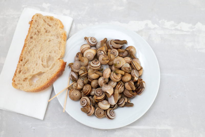 A white plate of escagot that was cooked in chicken stock, next to a single slice of bread