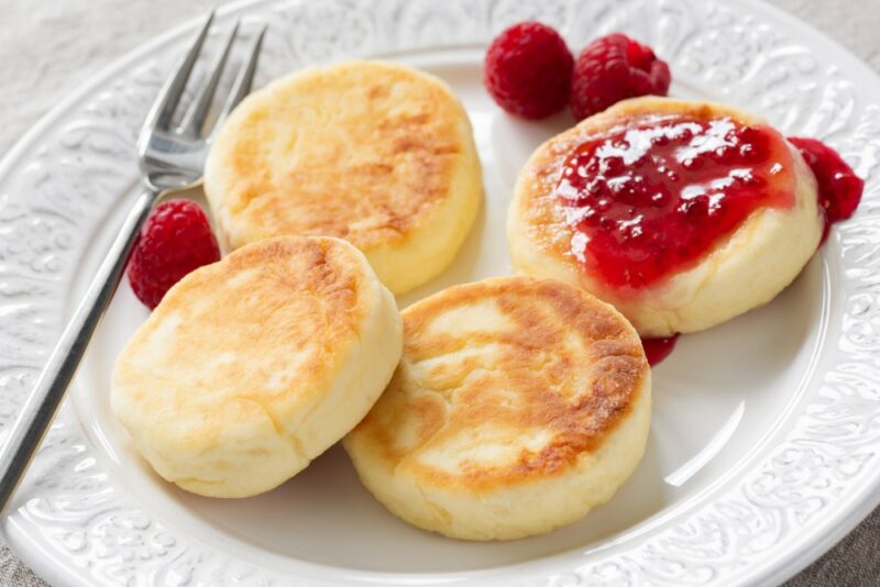 A white bowl containing four syrnikis. One is covered with raspberry jam and they are next to some raspberries and a fork