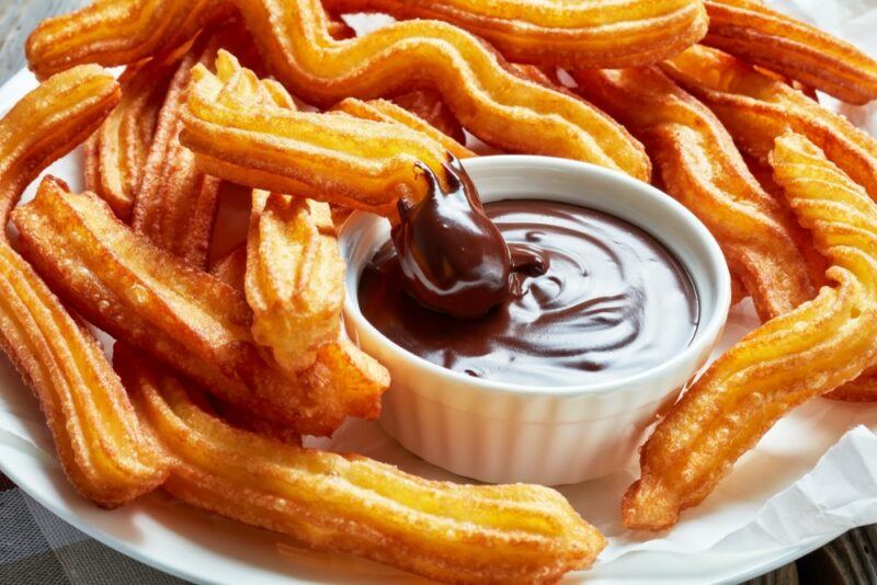 A white plate with many fried churros and a white bowl of chocolate sauce. One of the churros has been dipped in the sauce