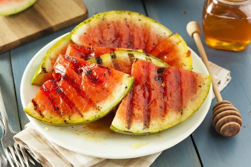 A white plate with grilled watermelon pieces, next to a stirrer for honey and a small jar of honey