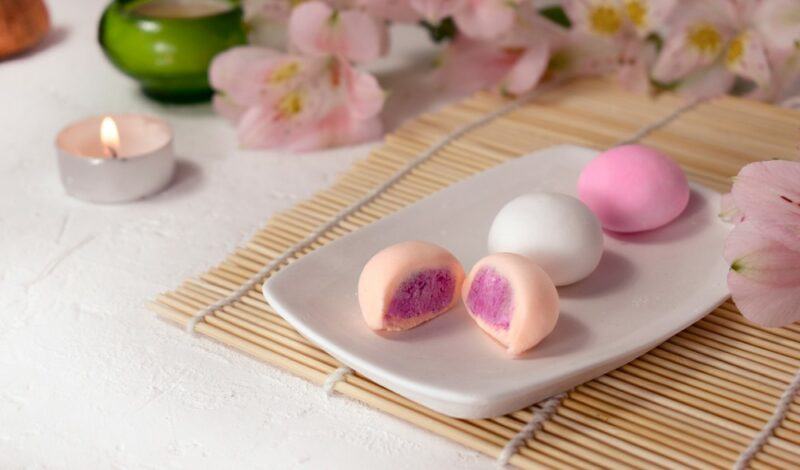 A bamboo mat with a white plate and pink and white pieces of mochi, plus pink flowers in the background