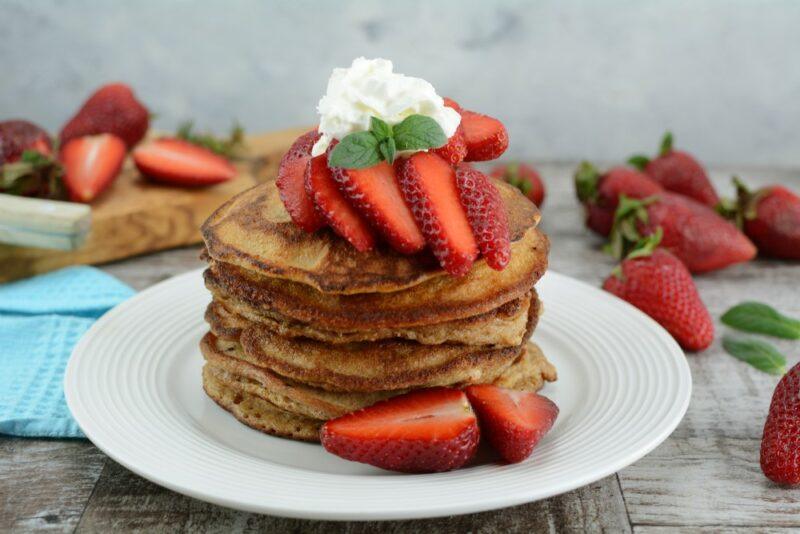 A white plate with a stack of protein pancakes with strawberries on top and various strawberries in the background