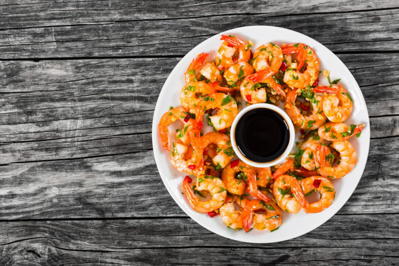 A white plate of shrimp on a wooden table with a small container of dipping sauce