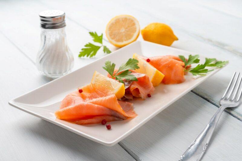 A white plate with smoked salmon, next to some greens, lemons, and salt