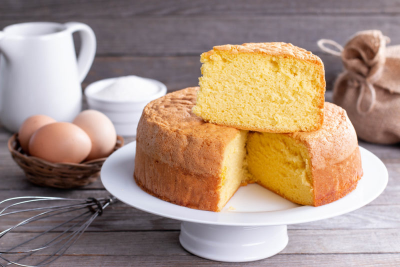 A white cake stand with a sponge cake where one piece has been cut out of the cake and placed on top