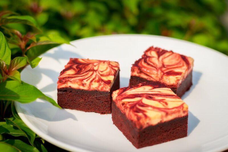 A white plate with three red velvet cream cheese brownies outside with leaves in the background