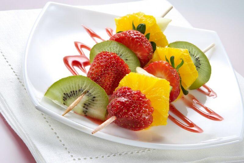 A square white plate with two fruit kebabs and fresh fruit sauce, the fruits include pineapple, kiwis, and strawberries