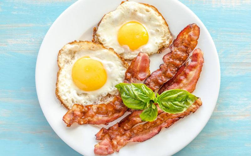 A white plate with two fried eggs, some strips of bacon and greens