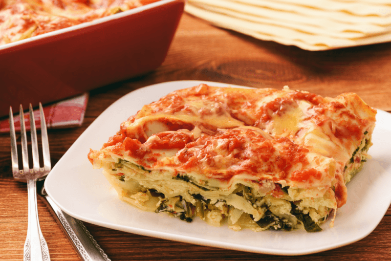 A white square plate with a large piece of vegetable lasagna, next to a fork.