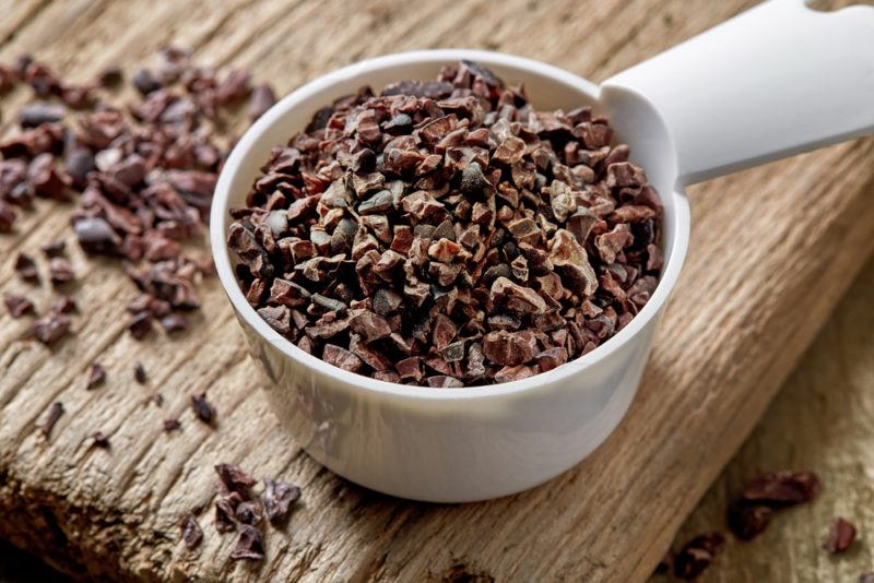 A white scoop that contains cacao nibs on a cutting board with more nibs