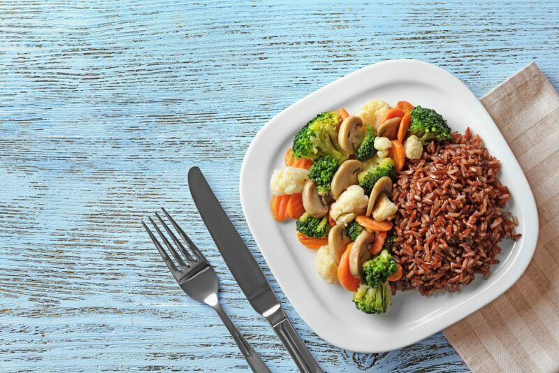 A white plate with a pile of brown rice and veggies, on a blue table next to a knife and fork