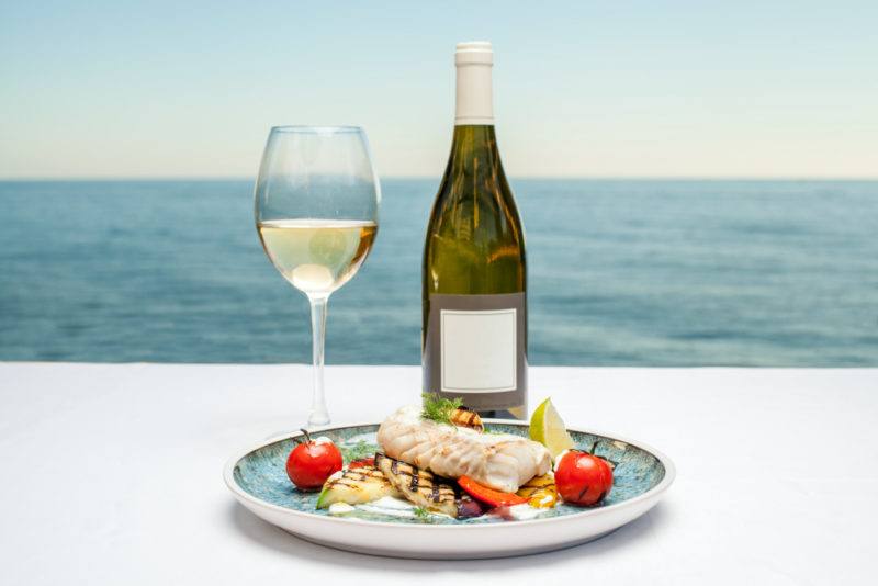 A glass of white wine and a bottle of wine next to a dish of cooked fish in front of the ocean