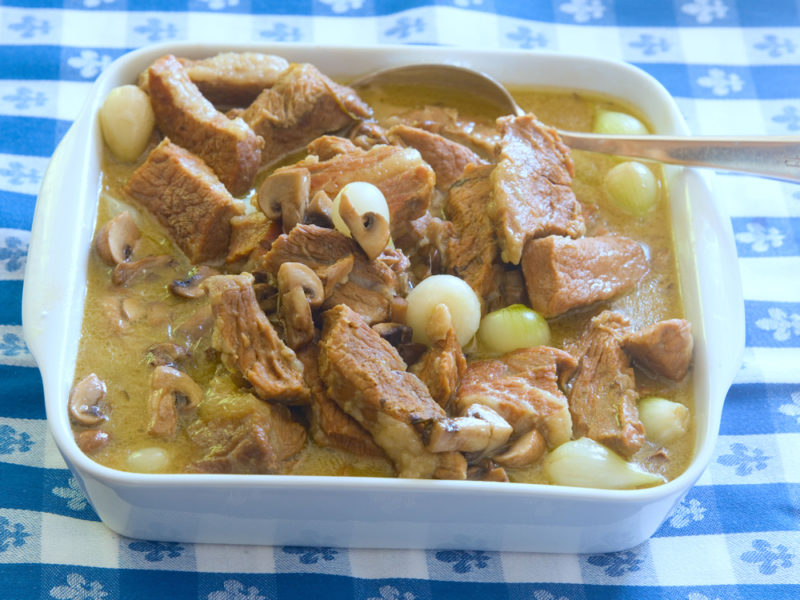 A white dish of veal stew with onions and a spoon