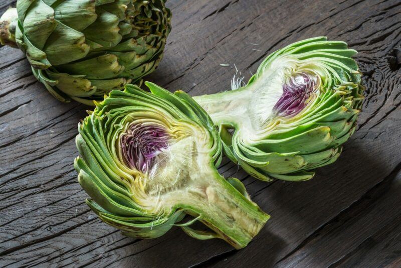 A whole artichoke and one that's been cut in half on a piece of wood.