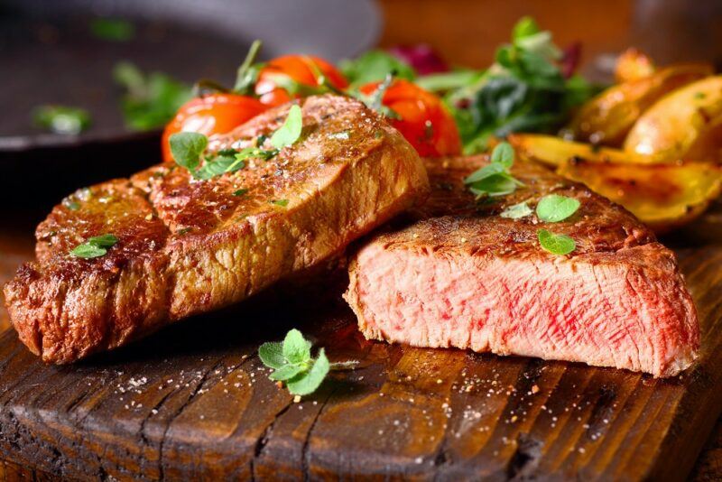 A wooden board with a whole piece of steak and half a steak, with some tomatoes and greens in the background