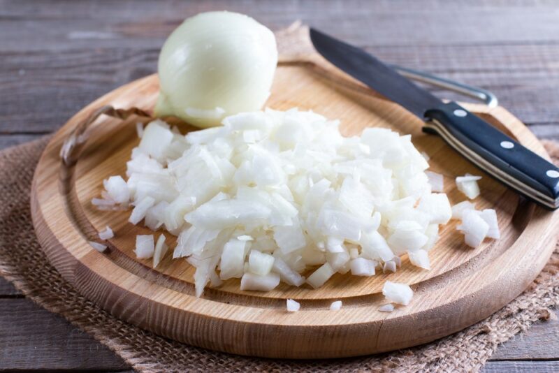 A round board with chopped raw white onion, a whole onion, and a knife