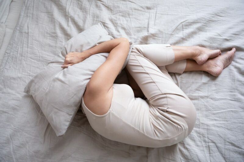 A woman on a white bed curled into a fetal position with her head under a pillow, crying