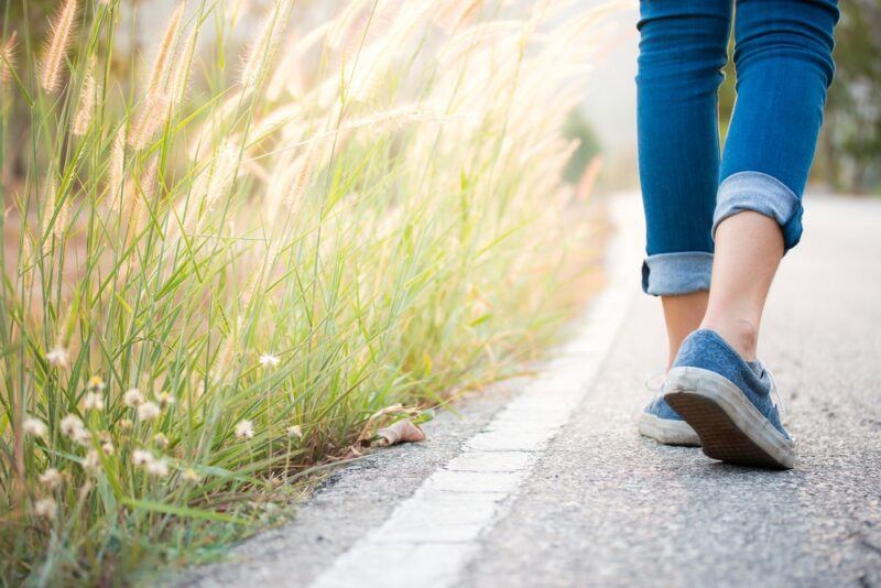 A young woman walking outside. You can see tall grass to her left, along with her shoes and jeans.