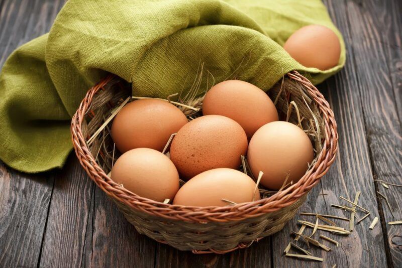 A basket on a wooden table, filled with eggs and some straw. There is also a green cloth on the table that houses a single egg.