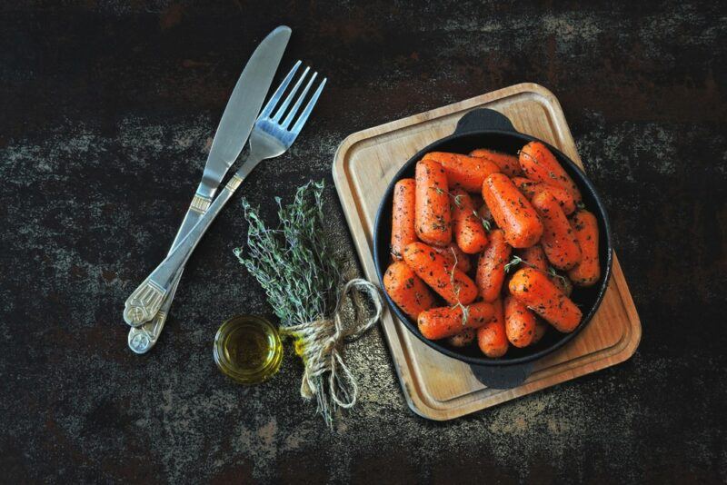 A wooden board with a black dish filled with cooked bagby carrots, some herbs, oil, a knife, and a fork