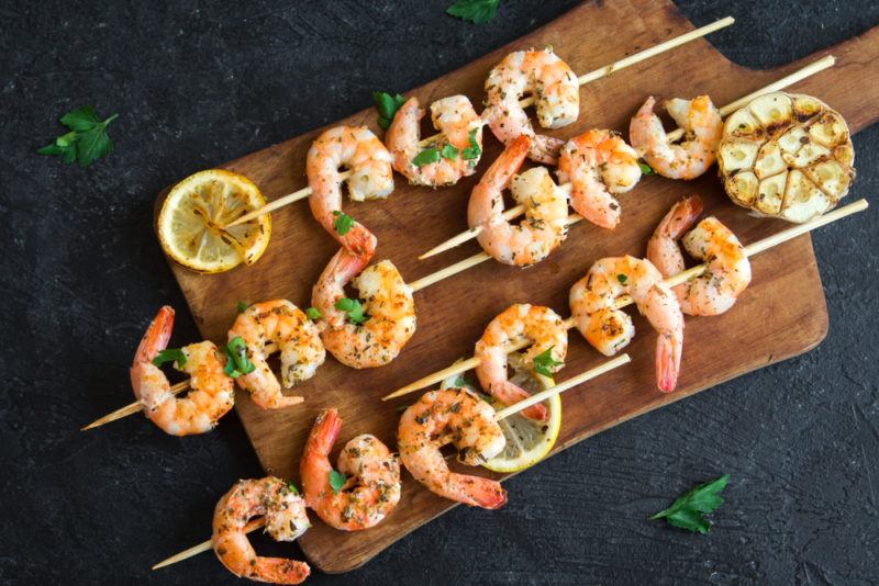 A wooden board against a black table, with shrimp skewers and slices of lemon