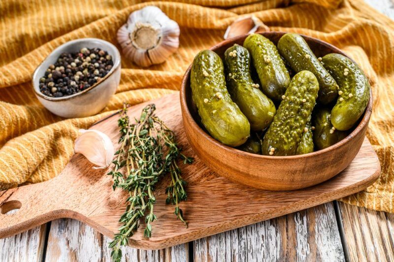 A wooden board with a bowl of pickles, next to some rosemary and garlic. There's also a yellow and brown cloth with a bowl of peppercorns and more garlic.