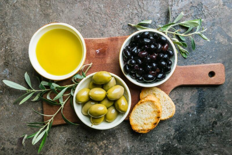 A wooden board with a bowl of green olives, a bowl of black olives, and a dish of olive oil. There are also some pieces of bread and olive branches